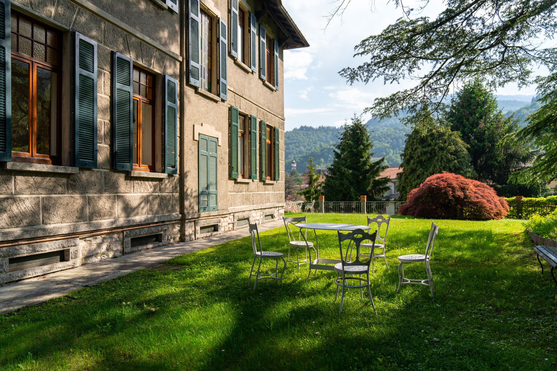Un bâtiment en pierre avec des volets verts, une pelouse verdoyante et une table entourée de chaises.