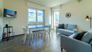 Una sala moderna con un sofá gris, una mesa de comedor con sillas, un televisor de pantalla plana montado en la pared y un amplio ventanal con vistas a la montaña.