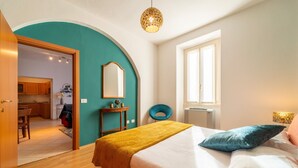 Un dormitorio con cama, escritorio, silla, ventana y puerta que da a una cocina.