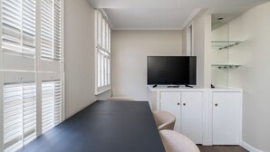 Quarto moderno com uma mesa escura comprida, uma TV de tela plana sobre um móvel branco e uma estante de vidro.
