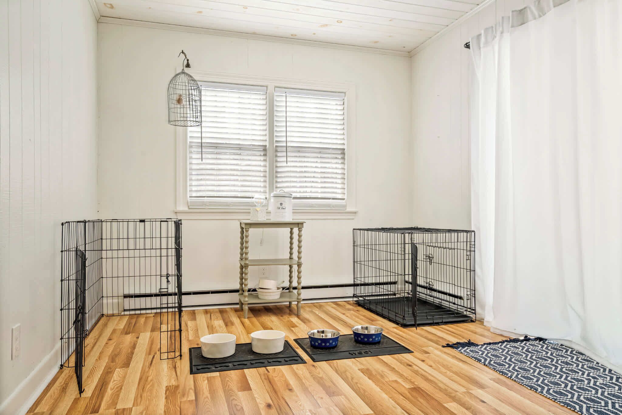 A room with two metal pet cages, a wooden table with bowls, and a window with blinds.