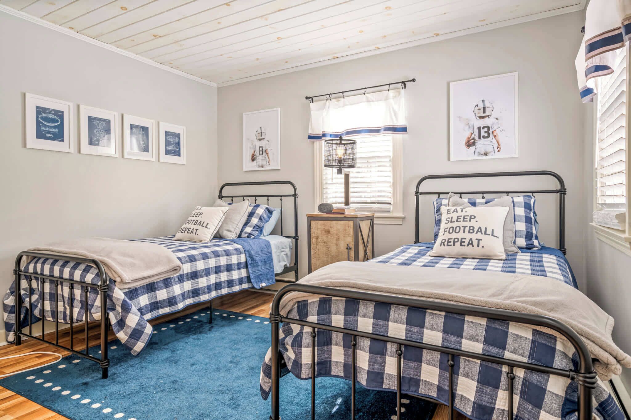 A bedroom with two metal beds, a blue and white checkered bedspread, a small wooden nightstand, and a window with blinds.