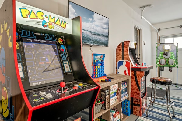 Une borne d’arcade Pac-Man dans une pièce où l’on peut voir un téléviseur diffusant une scène de plage, des étagères remplies de jeux et un petit tabouret.