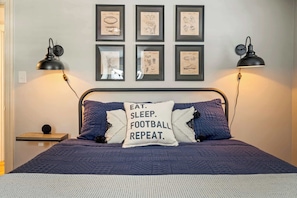 Un lit bien fait, avec un oreiller sur lequel on peut lire « MANGER. DORMIR. FOOTBALL. RÉPÉTER. » et une tête de lit. Deux lampes de bureau noires sont placées de chaque côté du lit, et six dessins encadrés ornent le mur au-dessus.