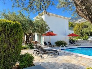 Un espace piscine avec des chaises longues et des parasols rouges, entouré de verdure et bordé d’un bâtiment blanc.