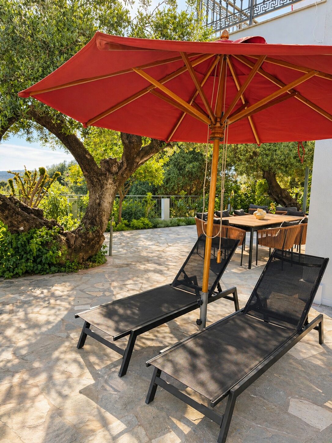 A patio with two lounge chairs, a red umbrella, and a table with cushions.
