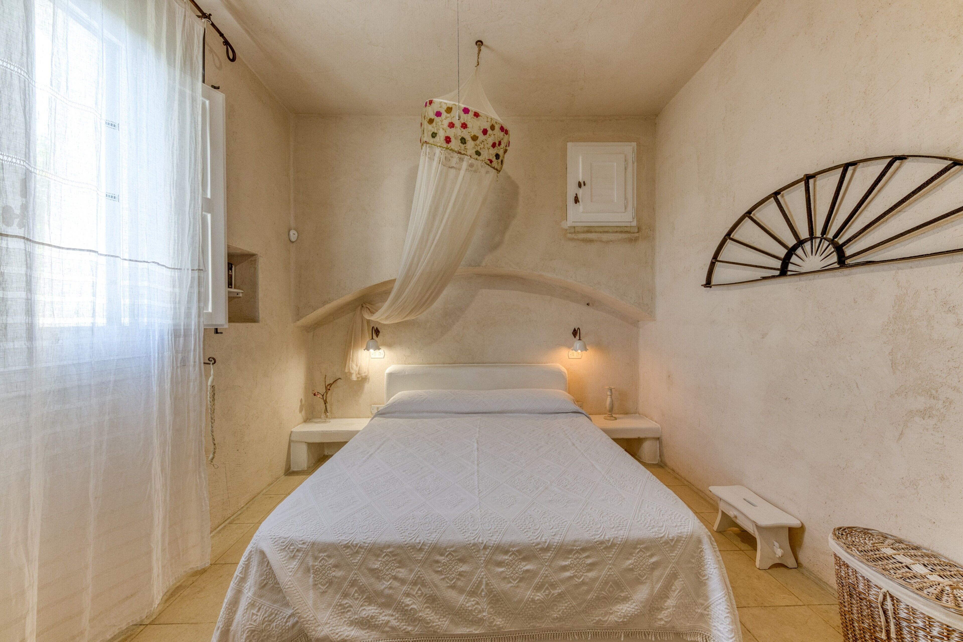 A bedroom with a bed, bedside tables, a bench, a hanging canopy, a wall-mounted fan, and a door.
