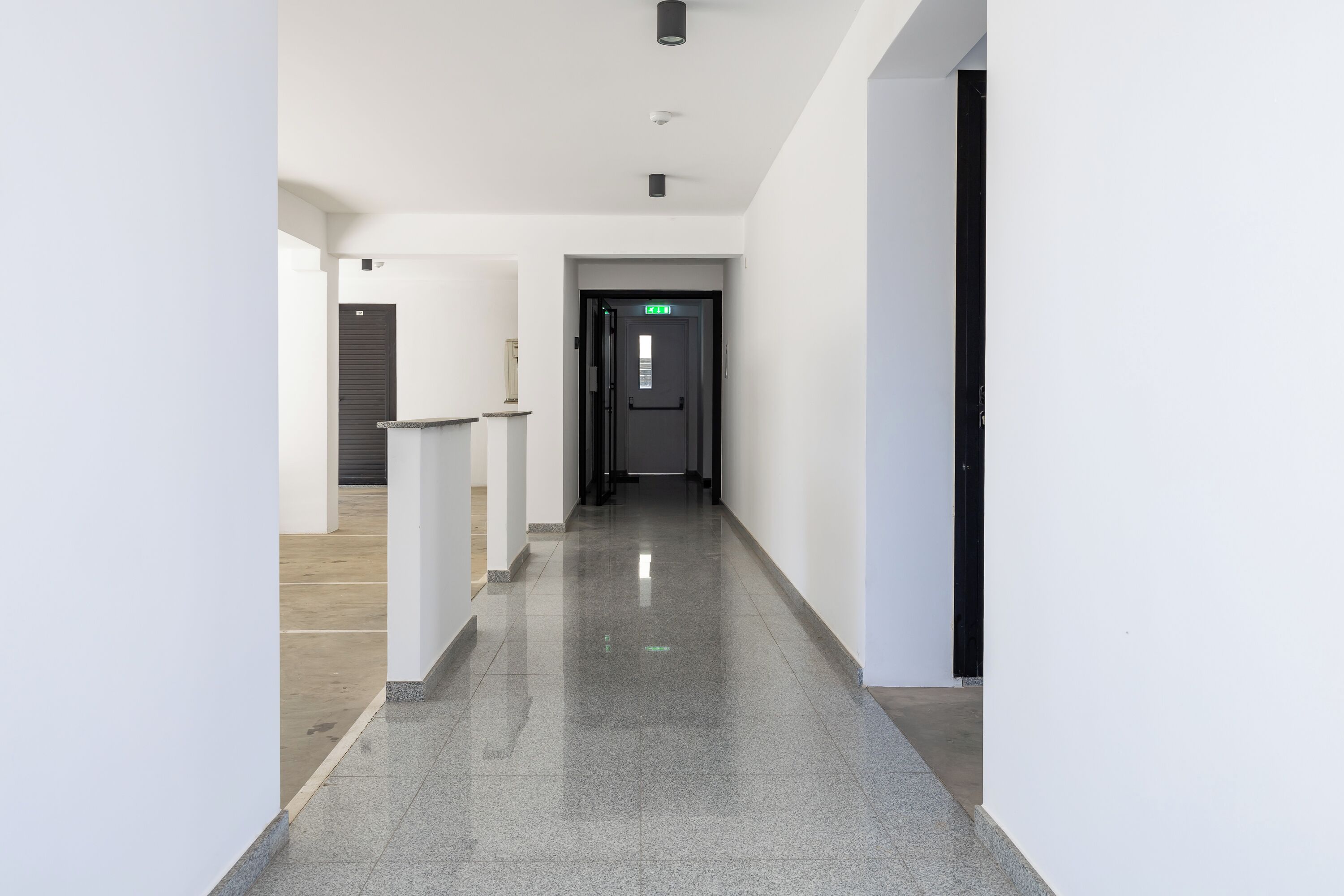 A modern, well-lit hallway with a polished floor, leading to a door with an emergency exit sign.