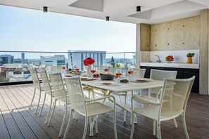 Eine Dachterrasse mit Esstisch für sechs Personen, umgeben von Korbstühlen, einer Küchenzeile mit Spüle und Arbeitsplatte sowie Blick auf die Stadt.
