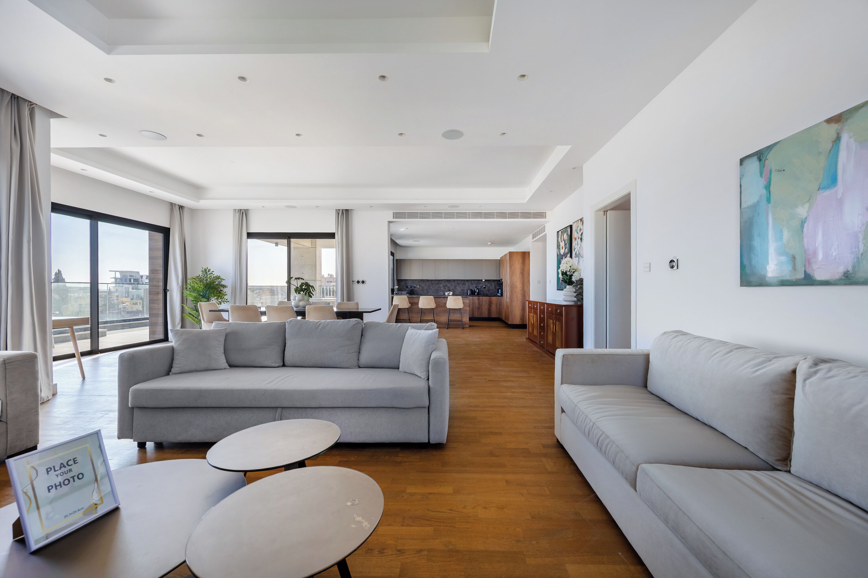 A spacious living area with a grey sofa, a coffee table, and a dining table. There is a kitchen in the background with modern appliances and a large window offering a view of the city.