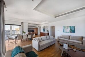 Ein geräumiger Wohnbereich mit Sofa, Couchtisch und Essstühlen. Im Hintergrund ist eine Küche erkennbar, und ein großes Fenster bietet Blick auf die Stadt.