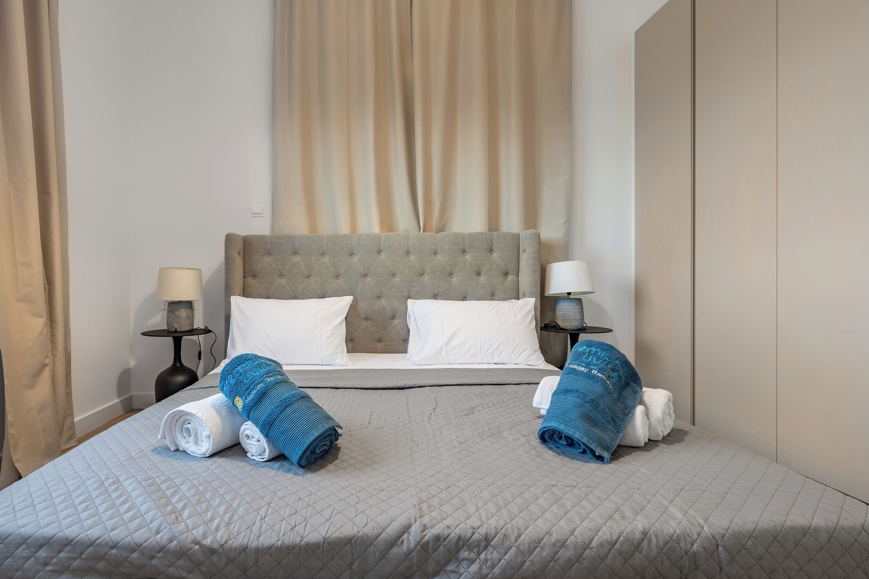 Een net opgemaakt bed met een grijze deken, twee opgerolde handdoeken en een blauwe handdoek.
