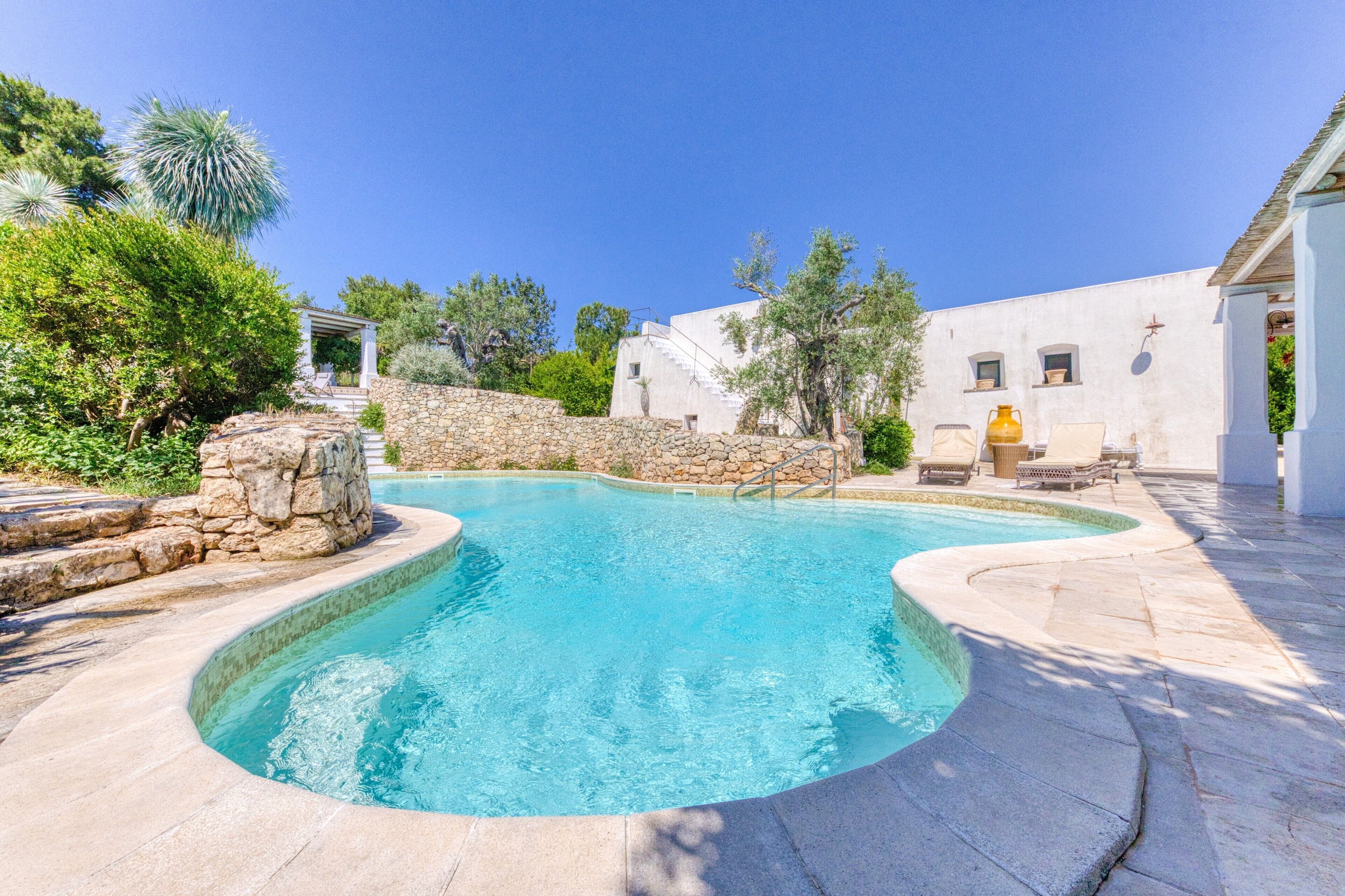 A swimming pool with stone steps, a white building, and surrounding greenery.