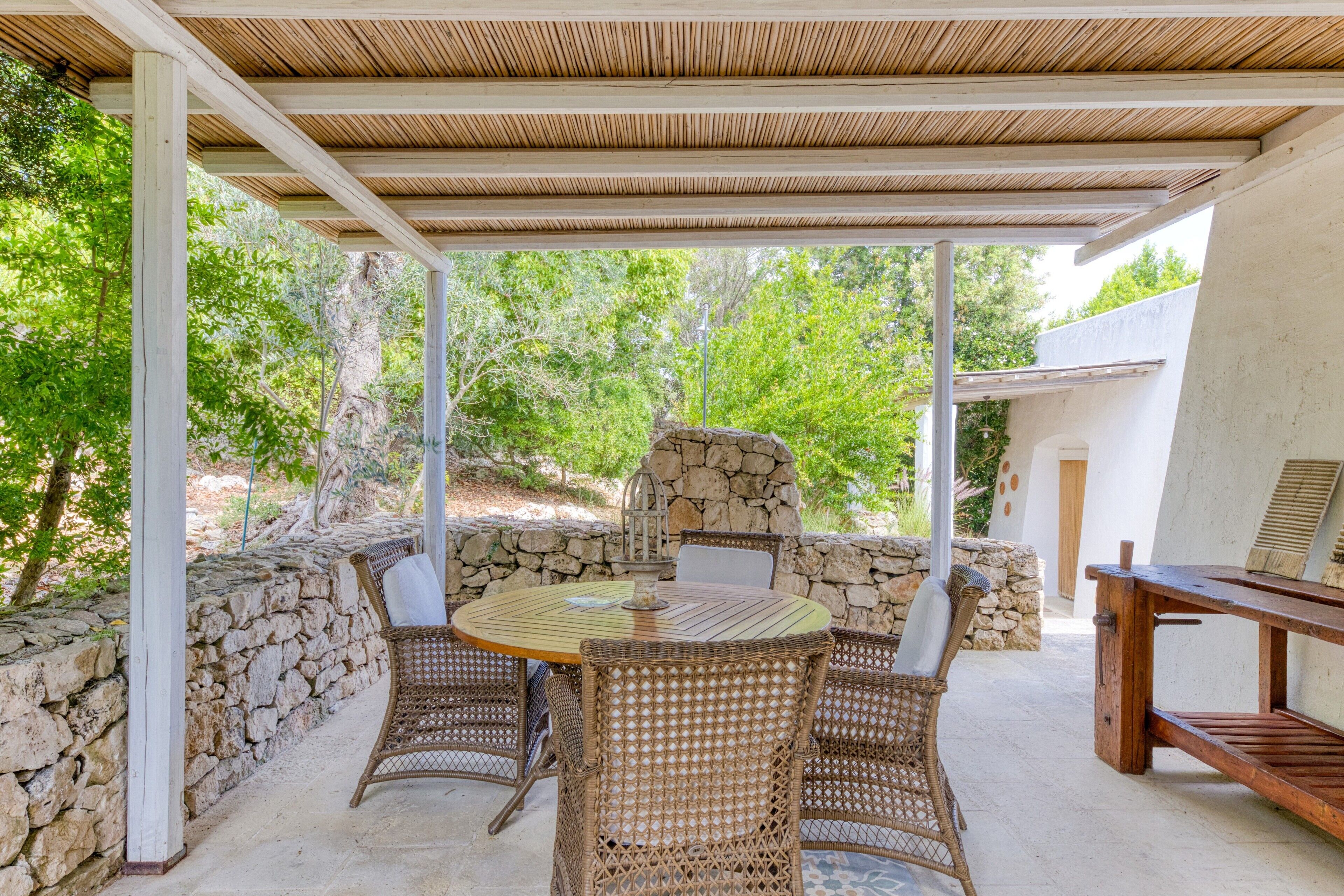 Eine Terrasse im Freien mit einem runden Tisch, Korbstühlen, einer Steinmauer und einer Pergola.