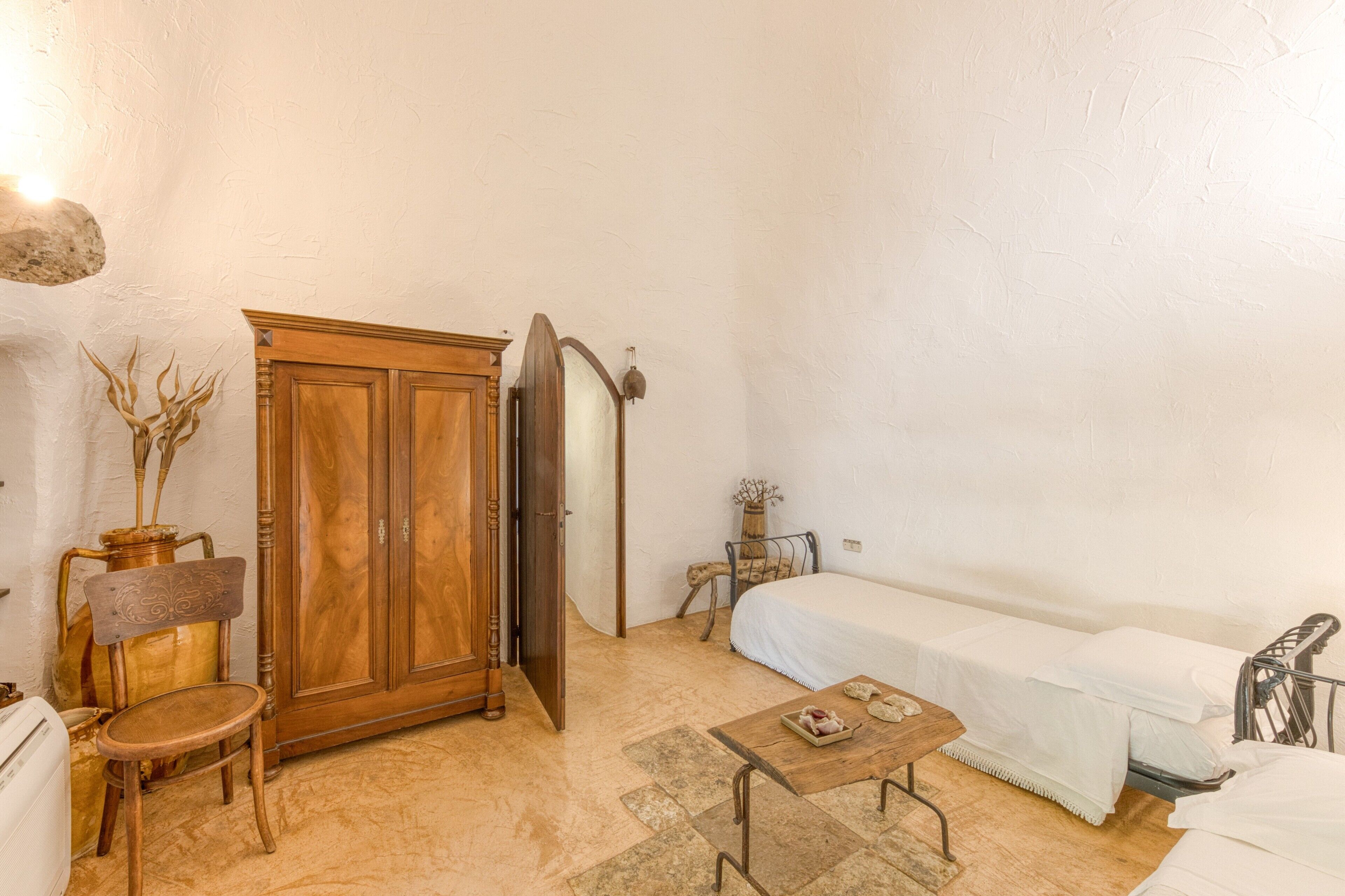Una habitación pequeña con un armario de madera, una cama, una silla, una mesita y una lámpara fijada a la pared.