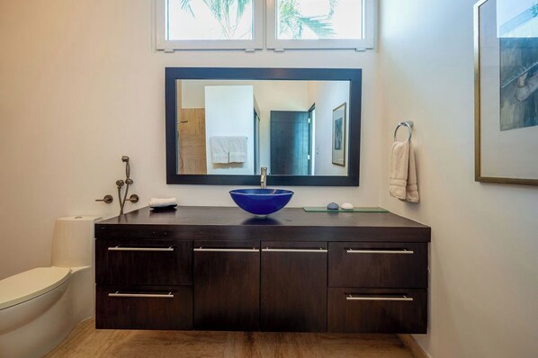 A bathroom with a dark wooden vanity, a blue basin, a mirror, a toilet, and a framed picture on the wall.