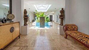 Un hall d’entrée spacieux, doté d’une verrière, d’un grand bac à plantes au centre, d’un banc en bois et d’une vitrine contenant des objets décoratifs.