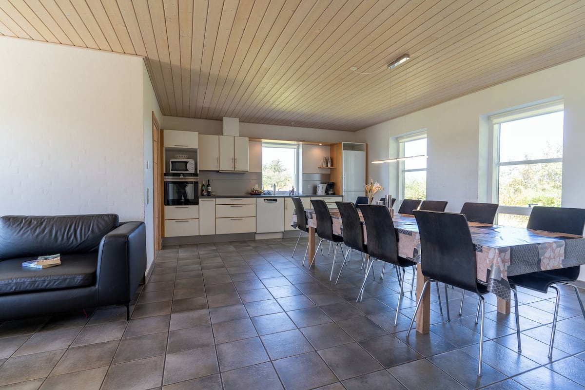 A modern kitchen with a dining area, featuring a black leather sofa, a dining table with chairs, and a large window providing natural light.