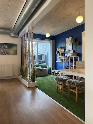 A modern interior with a wooden floor, a green artificial turf area, a white radiator, and a large window. - Den Gamle Smedje (Fjerritslev)