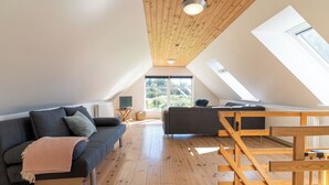 A spacious loft bedroom with a high ceiling, wooden beams, a skylight, a built-in bed, and a large window offering a view of the outdoors. - Den Gamle Smedje (Fjerritslev)