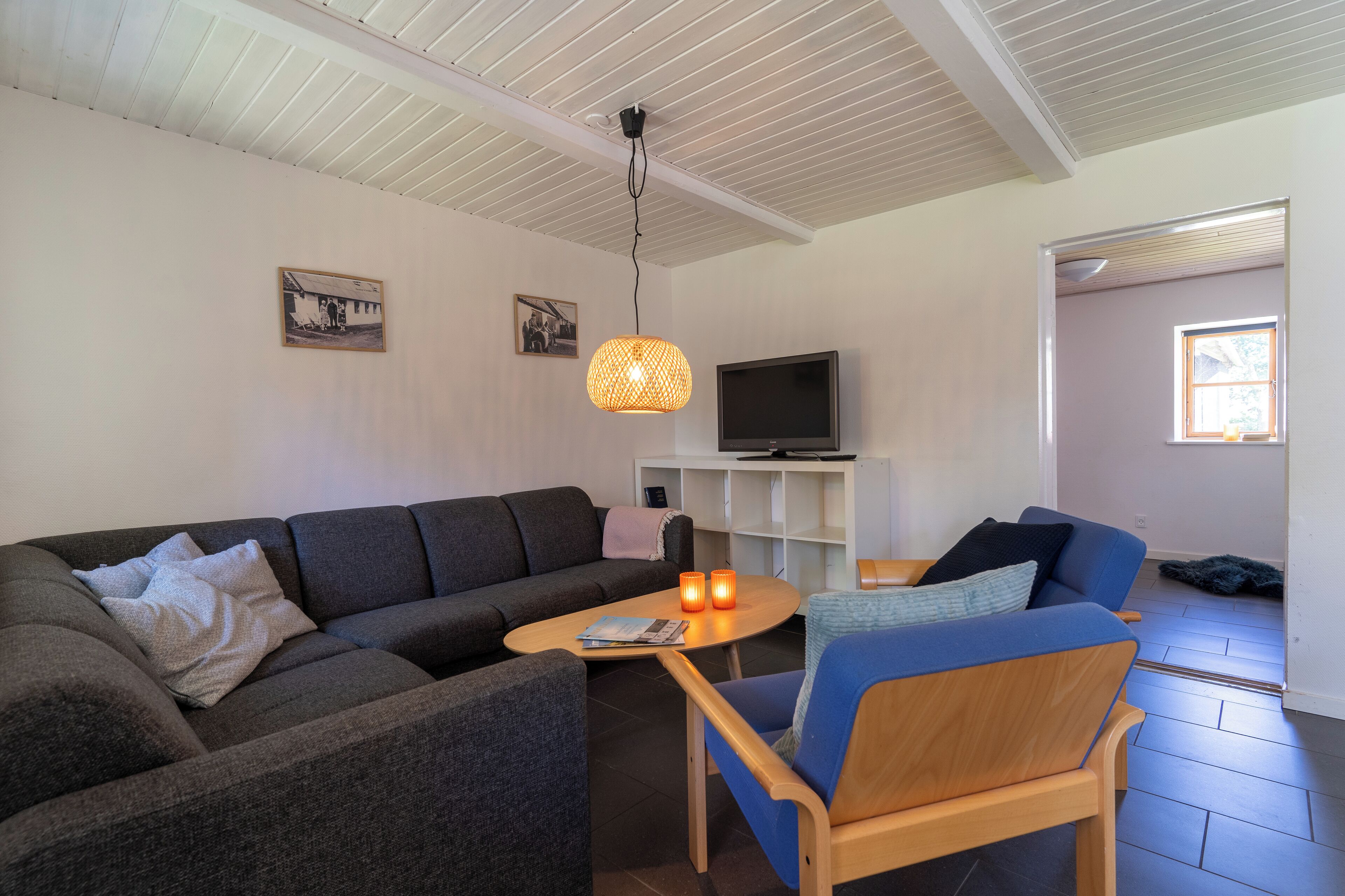 A living room with a sofa, a coffee table, a blue armchair, a television on a white stand, and a hanging light fixture.
