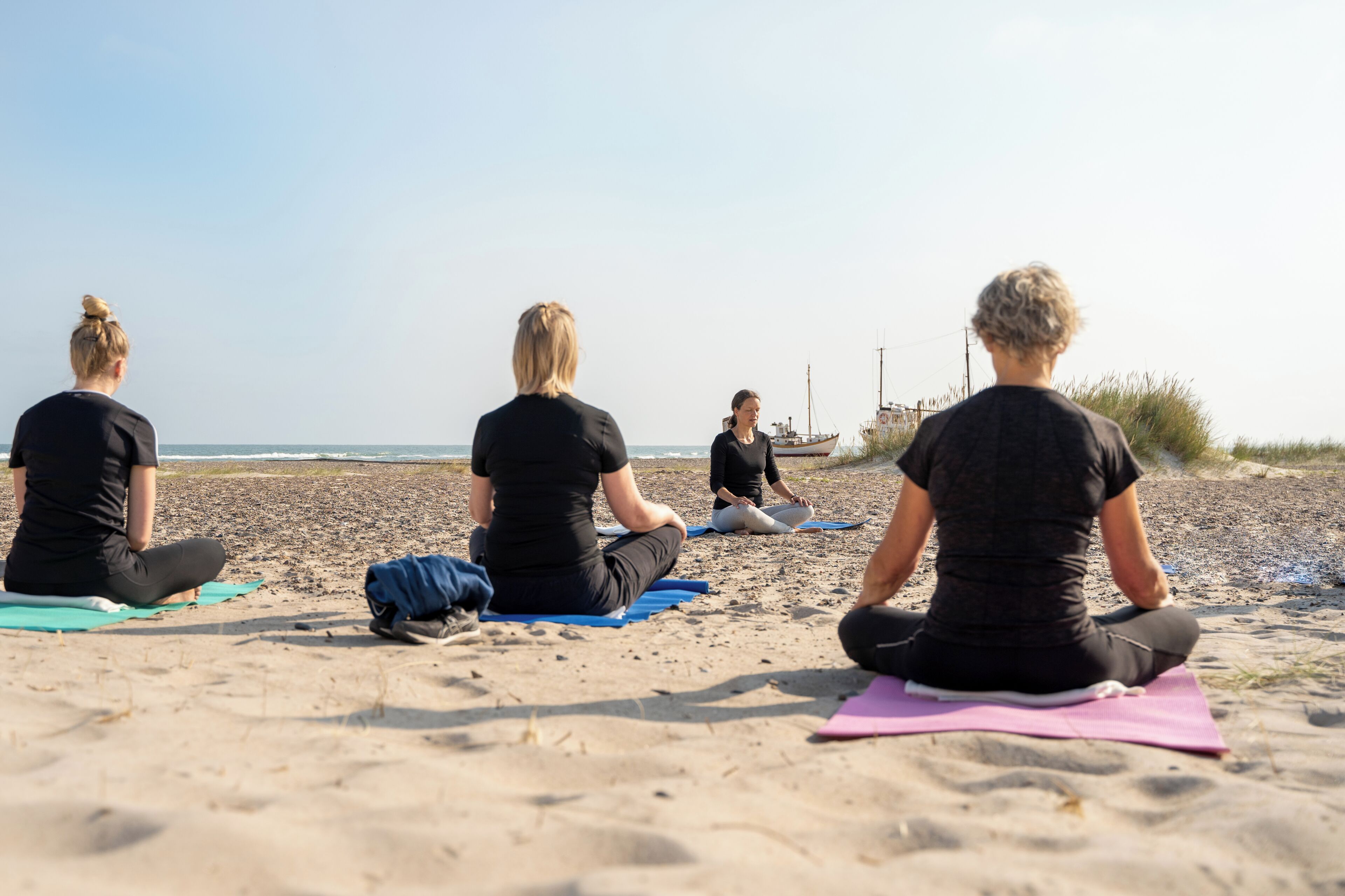 Vier personen zitten op yogamatten aan een zandstrand, met een boot op de achtergrond.