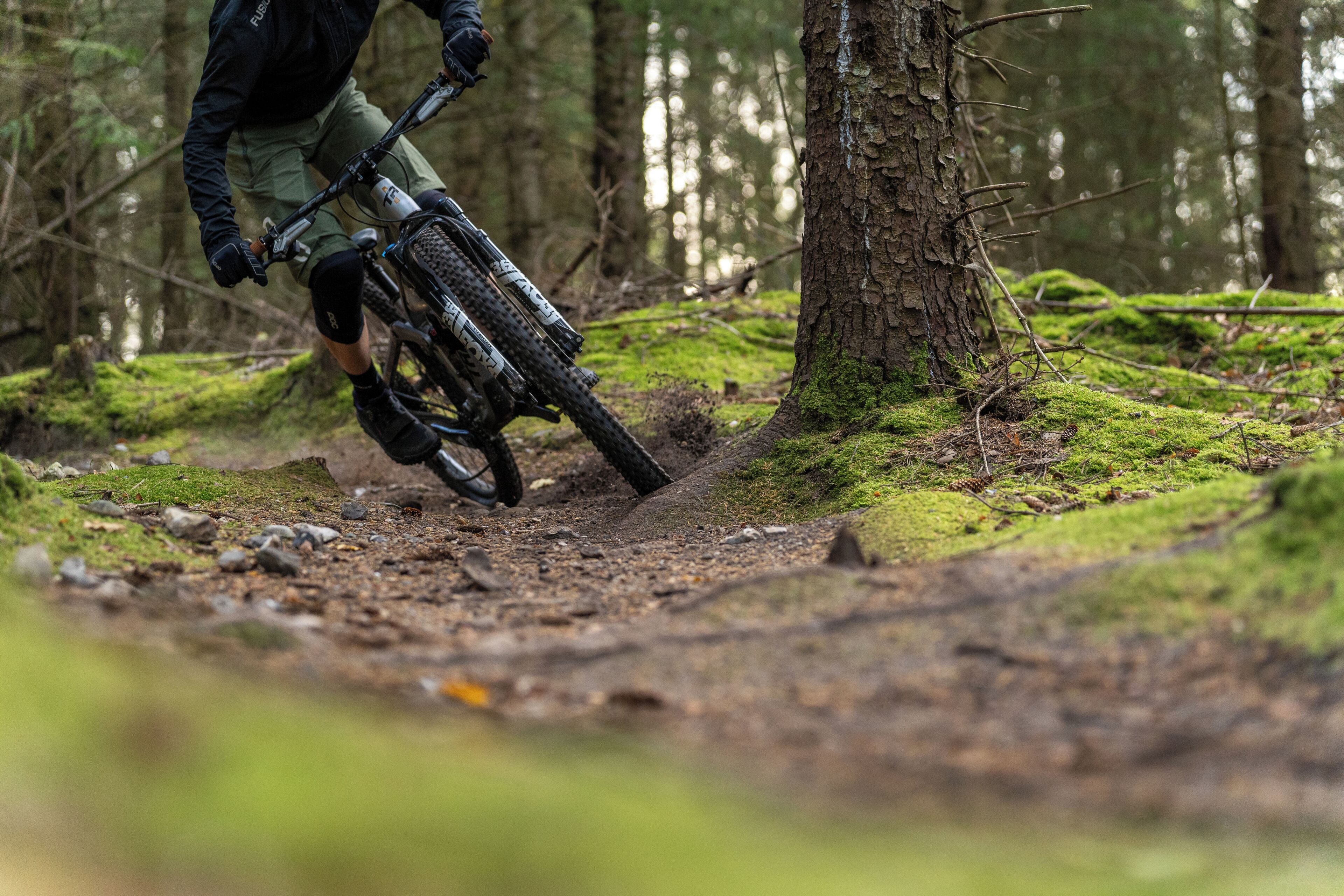 Una persona montando una bicicleta de montaña por un área boscosa con árboles y un sendero de tierra.