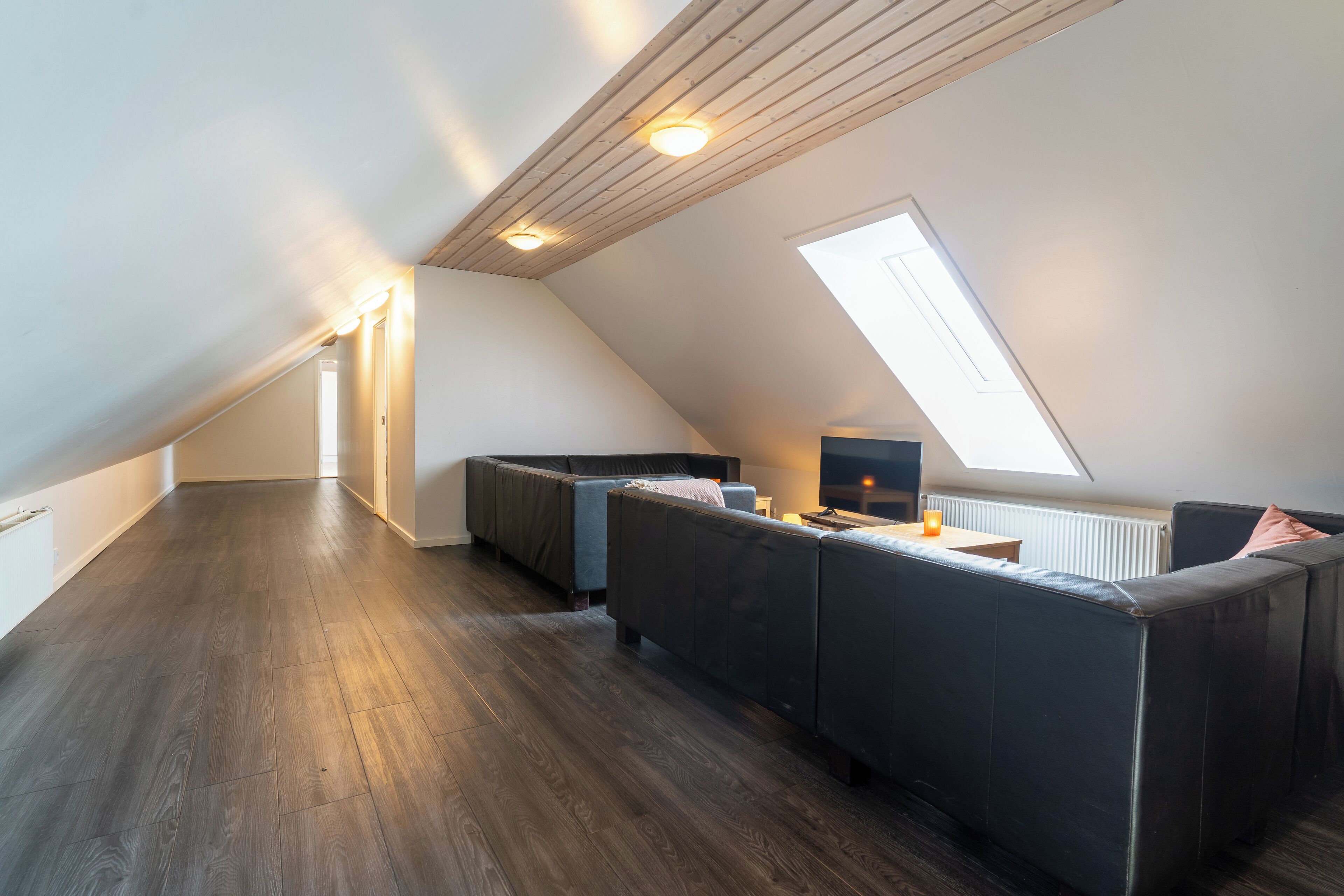 Un loft moderno con techo inclinado, vigas de madera, tragaluz y piso de madera oscura.