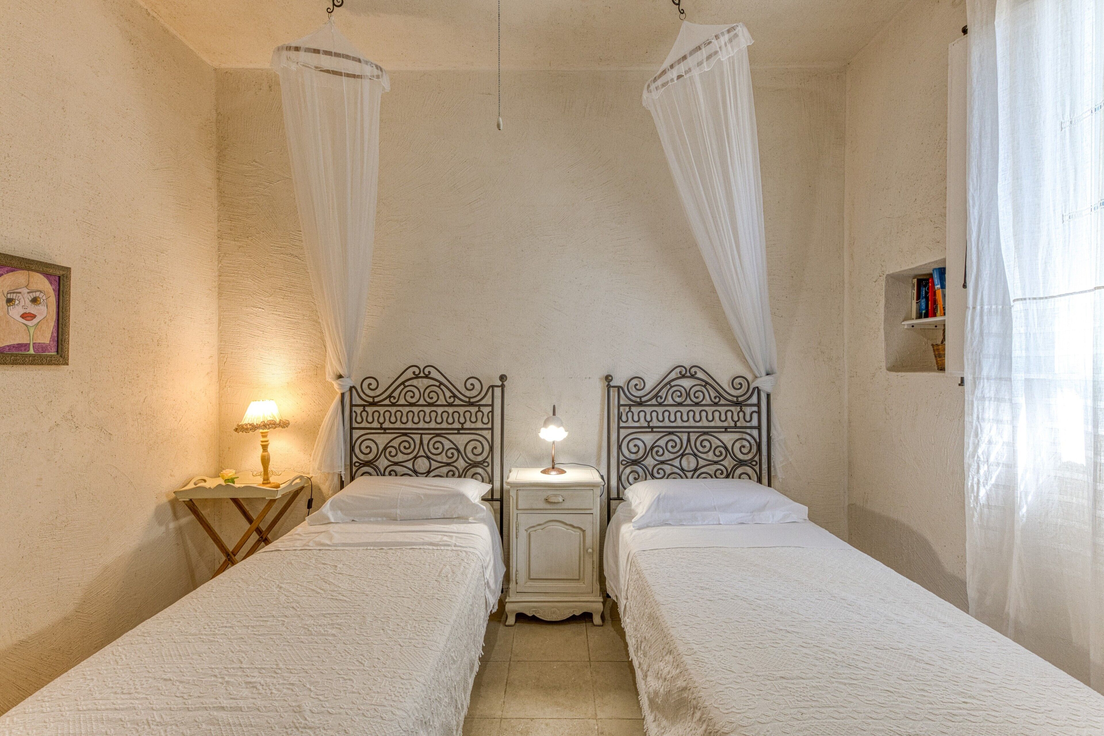 A room with two beds, a bedside table with a lamp, a wall-mounted shelf with books, and a canopy bed.