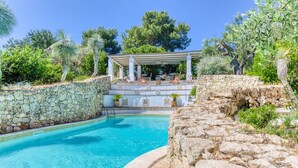 Una piscina con un muro de piedra, un espacio de descanso cubierto y vegetación circundante.