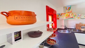 Una cocina con una olla naranja grande etiquetada 'CUCINIAMO', una puerta roja y un cuadro abstracto y colorido sobre el fregadero.