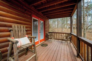 A wooden deck with a bench and two wicker chairs, a wooden door with a red frame, and a view of a forested area. - Kodiak Bear-hot Tub-playground-location! (Broken Bow)