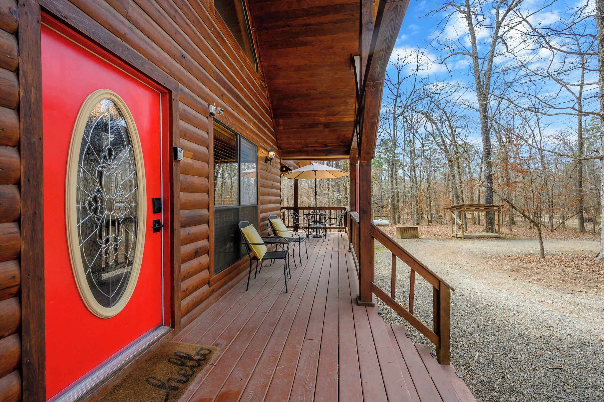 A log cabin with a red door, a covered porch with outdoor seating, and a view of the surrounding forest.