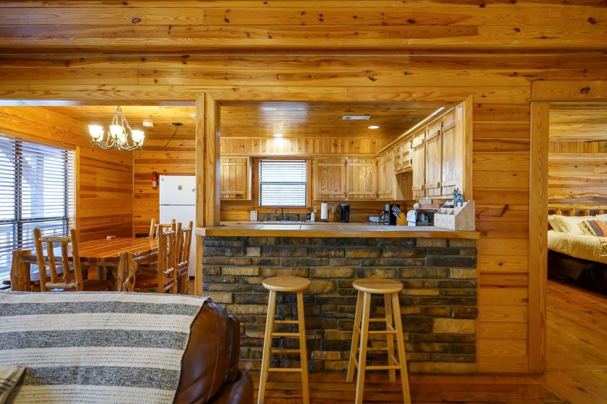 Een gezellig chalet-achtig vertrek met houten inrichting, een accentmuur van steen, een eethoek met tafel en stoelen, en een kitchenette met een bar.