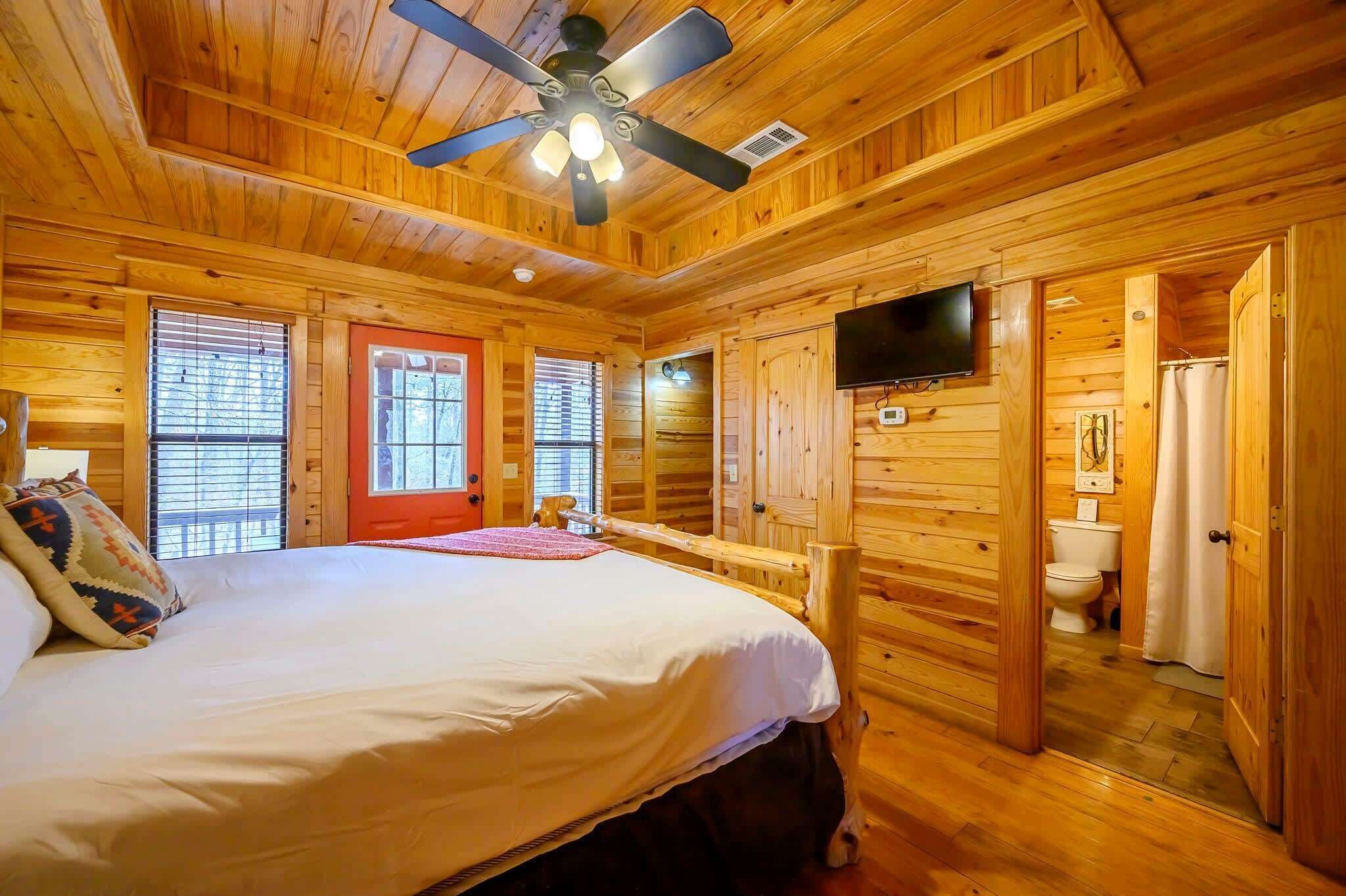 A wooden cabin bedroom with a bed, a ceiling fan, a television, and a bathroom visible through an open door.