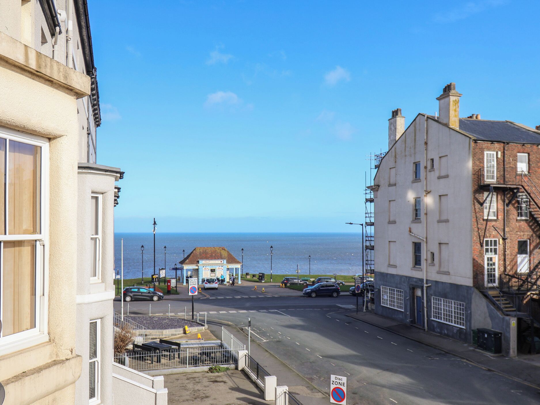 Rua costeira com edifícios, placa de proibido estacionar e vista para o mar.