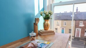 Un piano di lavoro in cucina con un barattolo di biscotti al tè, un set di cucchiai di legno, un vaso con fiori bianchi e una finestra vista edifici.