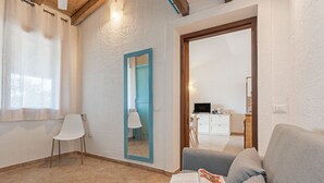 Una sala de estar moderna con un sofá gris, una silla blanca y un espejo con marco azul. Hay un libro sobre el sofá, y se ve una cocina a través de una puerta abierta.
