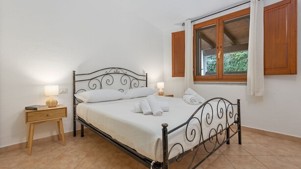 Un dormitorio con estructura de cama metálica, ropa de cama blanca, una mesita de noche con lámpara y una ventana con contraventanas de madera.