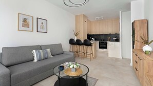 En moderne stue med en grå sofa, et glasbord og et køkkenområde med træskabe og sorte stole.