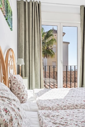 Une chambre à coucher avec un lit, une literie à motifs floraux, une table de chevet avec une lampe, et une vue sur un balcon agrémenté d’un palmier.