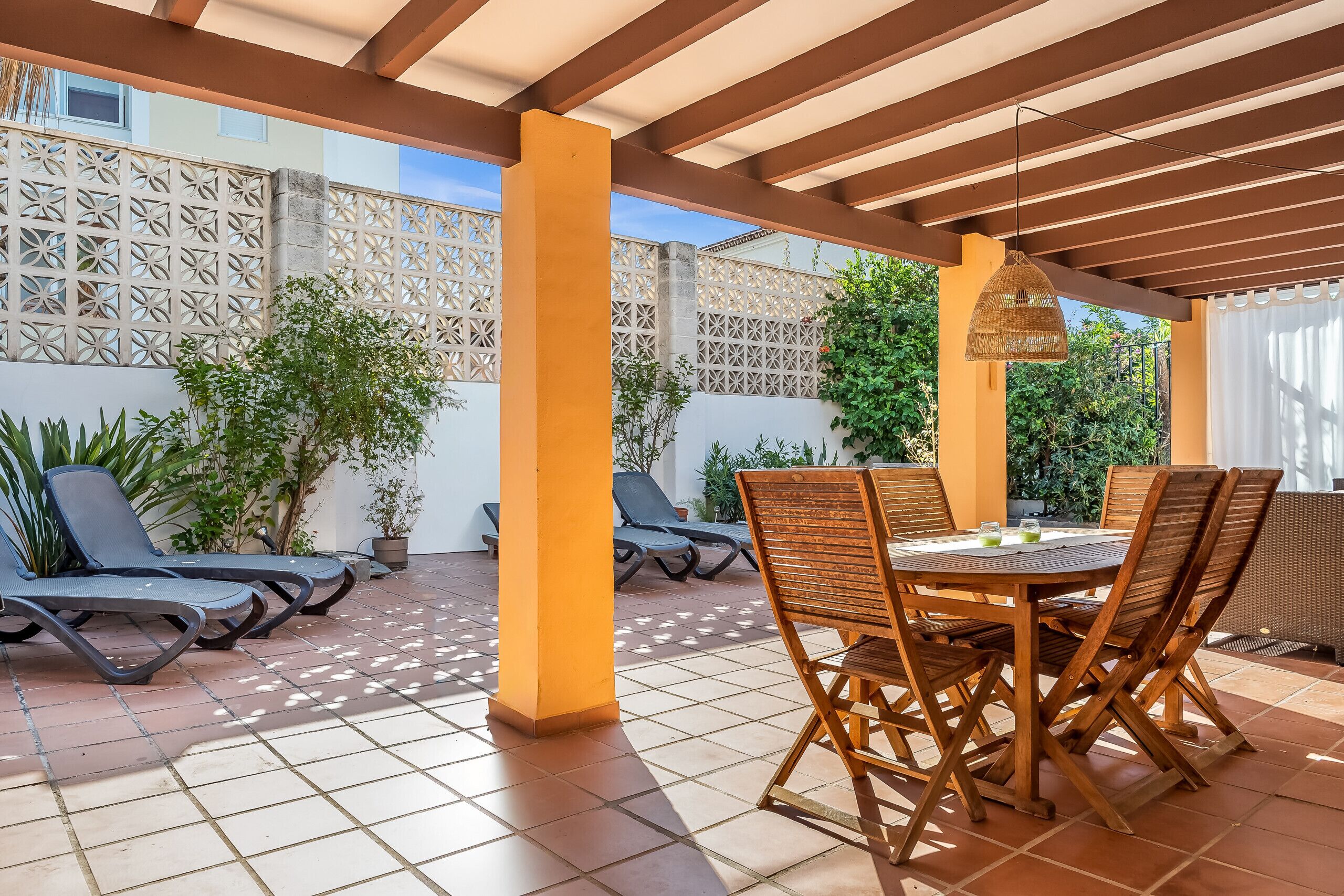 Un patio techado al aire libre con mesa y sillas, sillones de sol y una valla decorativa.
