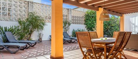 Un patio techado al aire libre con mesa y sillas, sillones de sol y una valla decorativa.