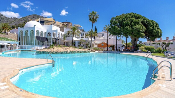 A large outdoor swimming pool with a wooden deck, surrounded by palm trees and a building with arched windows. - Charming Holiday Apartment in Nerja (Nerja)