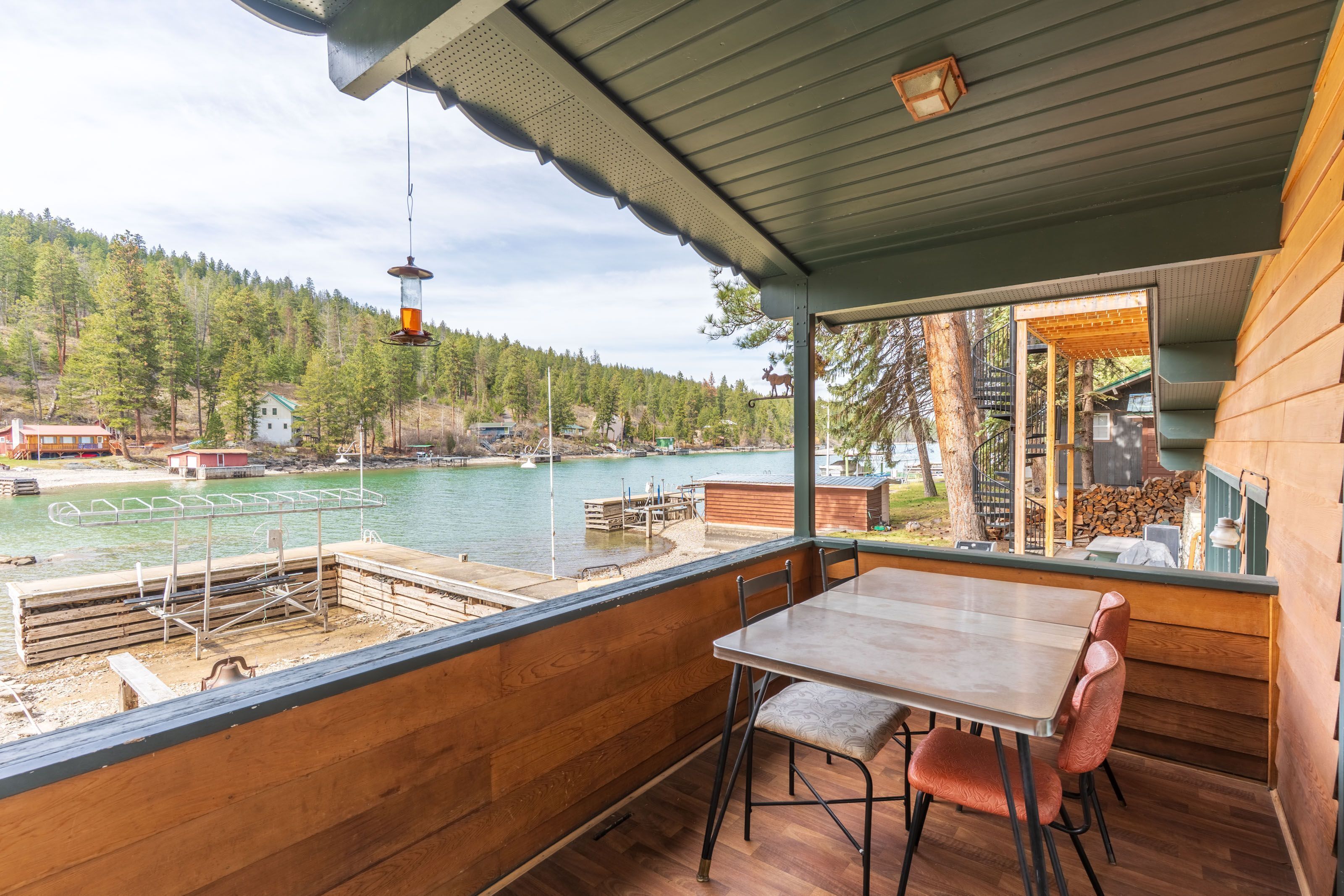 Une terrasse en bois avec une table et des chaises donnant sur un lac, d’où l’on peut admirer une colline boisée et, au loin, un quai.