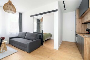 Un apartamento moderno con un sofá gris, una peque ña mesa redonda, una zona de cocina y una puerta corredera que da a un dormitorio.