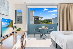 A hotel room with a bed, a desk with a computer, a small table with a bottle and glasses, and a balcony with a view of buildings and greenery. - Refreshed Esplanade Studio for Two (Urangan)