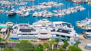 A marina with numerous boats docked, a modern building with solar panels, and a tropical landscape with palm trees. - Relaxed Esplanade Living near Pier & Marina (Urangan)