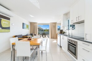 A modern kitchen with white cabinets, stainless steel appliances, and a dining area with a wooden table and white chairs. - Relaxed Esplanade Living near Pier & Marina (Urangan)