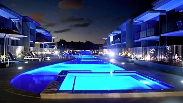 A swimming pool with blue lighting, surrounded by modern buildings with balconies and outdoor seating areas. - Relaxed Esplanade Living near Pier & Marina (Urangan)
